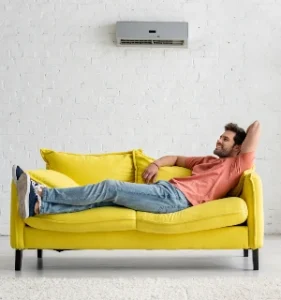 "Smiling man relaxing on a yellow sofa under an air conditioning unit, enjoying the cool air in a comfortable living space."