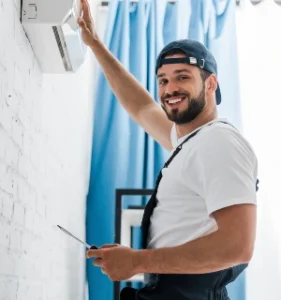 "Smiling technician repairing an air conditioning unit on a white brick wall, holding a screwdriver and looking at the camera, offering HVAC repair services."