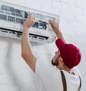 "Young repairman in a red cap replacing the filter of an air conditioning unit on a white brick wall, demonstrating air conditioning maintenance services."
